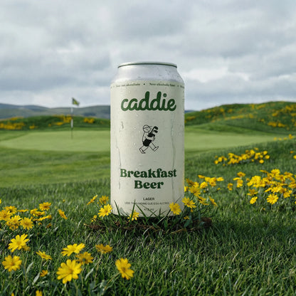Caddie Breakfast Beer can on a grassy field with yellow flowers and a cloudy sky.
